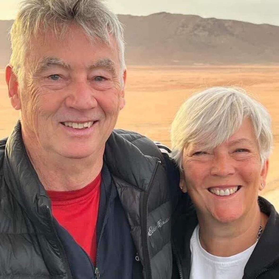 An older couple smiles while in front of mountains.