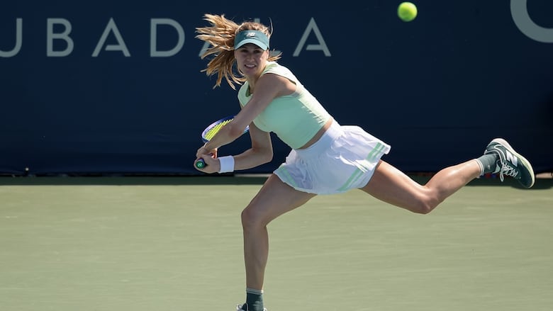 A female tennis player lunges to her right while returning a shot during an outdoor match on a hard court during the day.