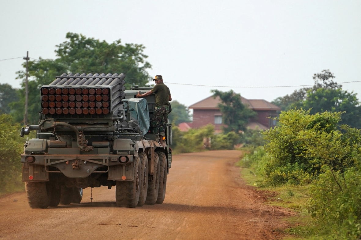 A soldiers rides atop a moving military vehicle.