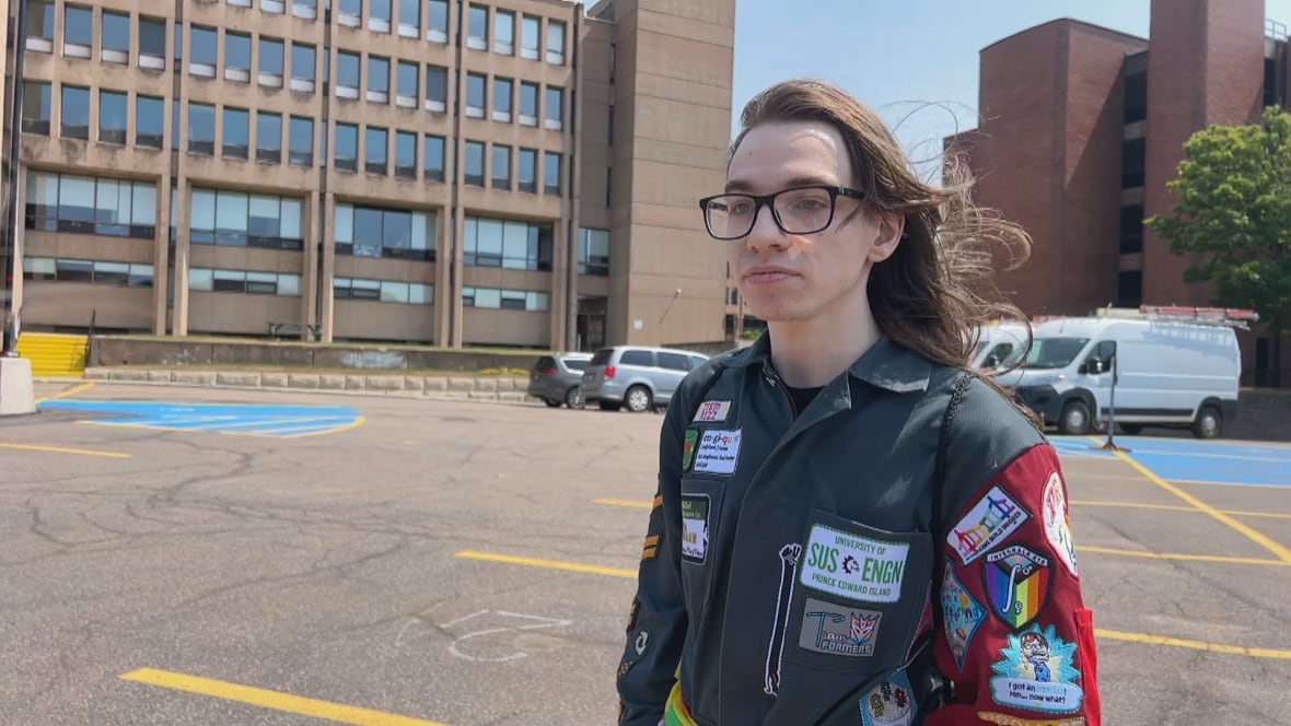 A student wearing a custom jacket with many patches stands in a parking lot.
