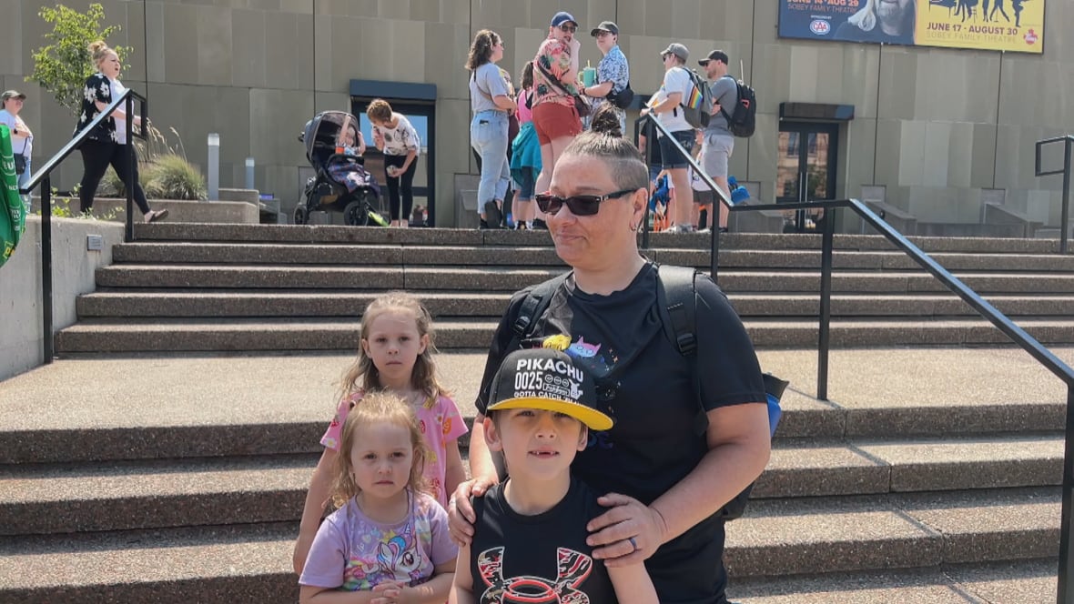 A woman wearing a black t-shirt and sunglasses stands behind her three kids.