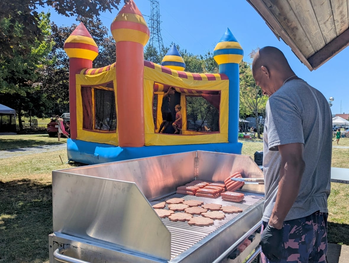 Picture from the side of a man grilling hot dogs and hamburgers on a bbq. Bouncy castle in the background.