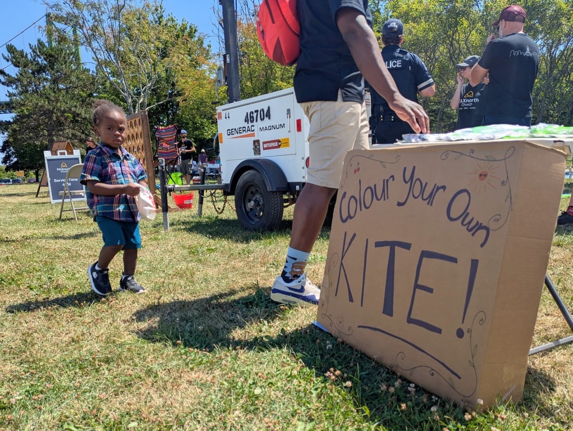 A kid looks at the camera while walking behind his dad. In the forefront there is a cardboard sign that says "colour your own kite."