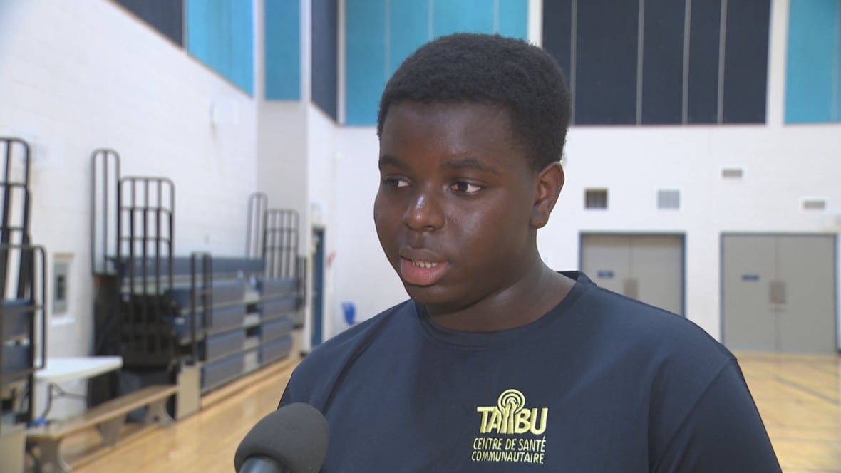 A black teenager in a cotton t shirt speaks into a reporter's microphone inside a school gymnasium. The gym is empty behind him
