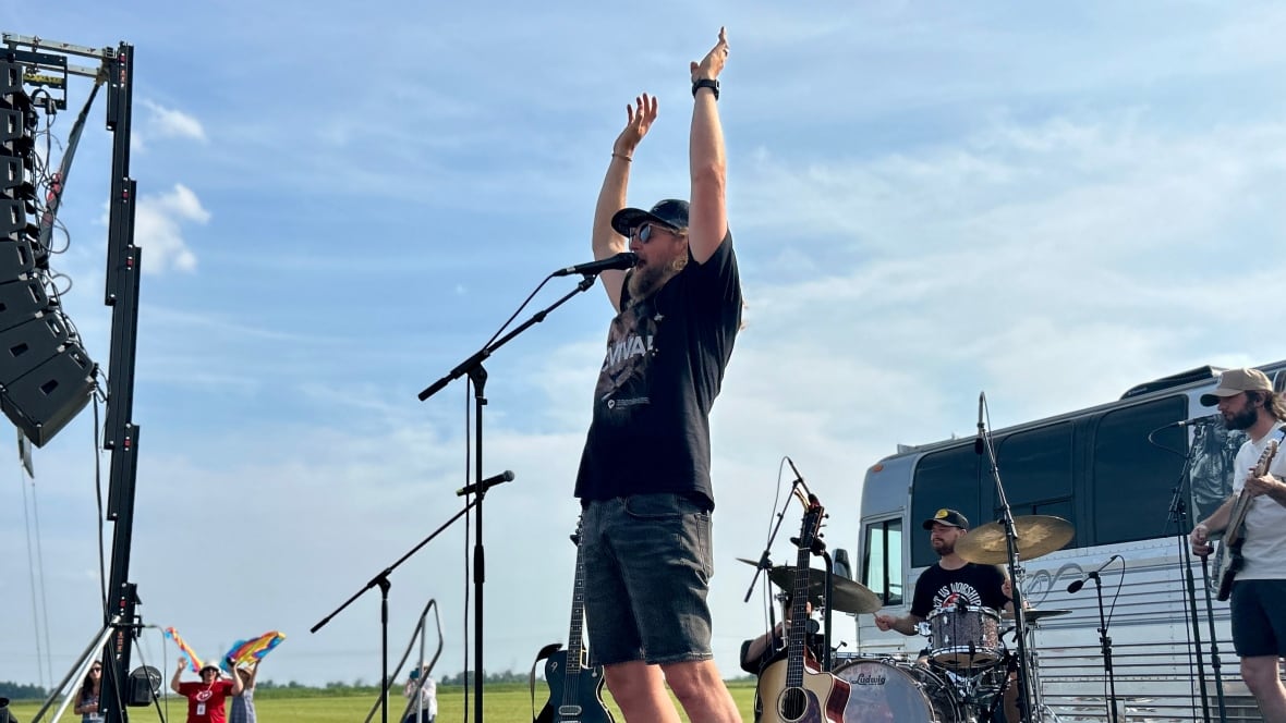 Un hombre con una camiseta, pantalones cortos y gafas de sol, sostiene sus manos sobre su cabeza en el escenario abierto sobre su cabeza, el baterista y guitarrista se presenta detrás de él.