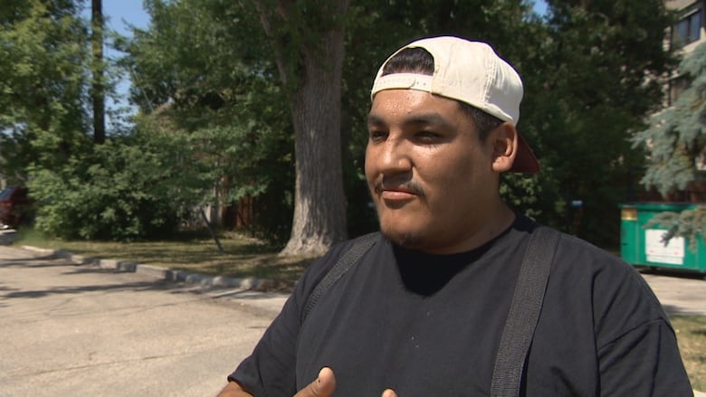 Man wearing white baseball cap turned backwards and black t-shirt stands on a residential street.