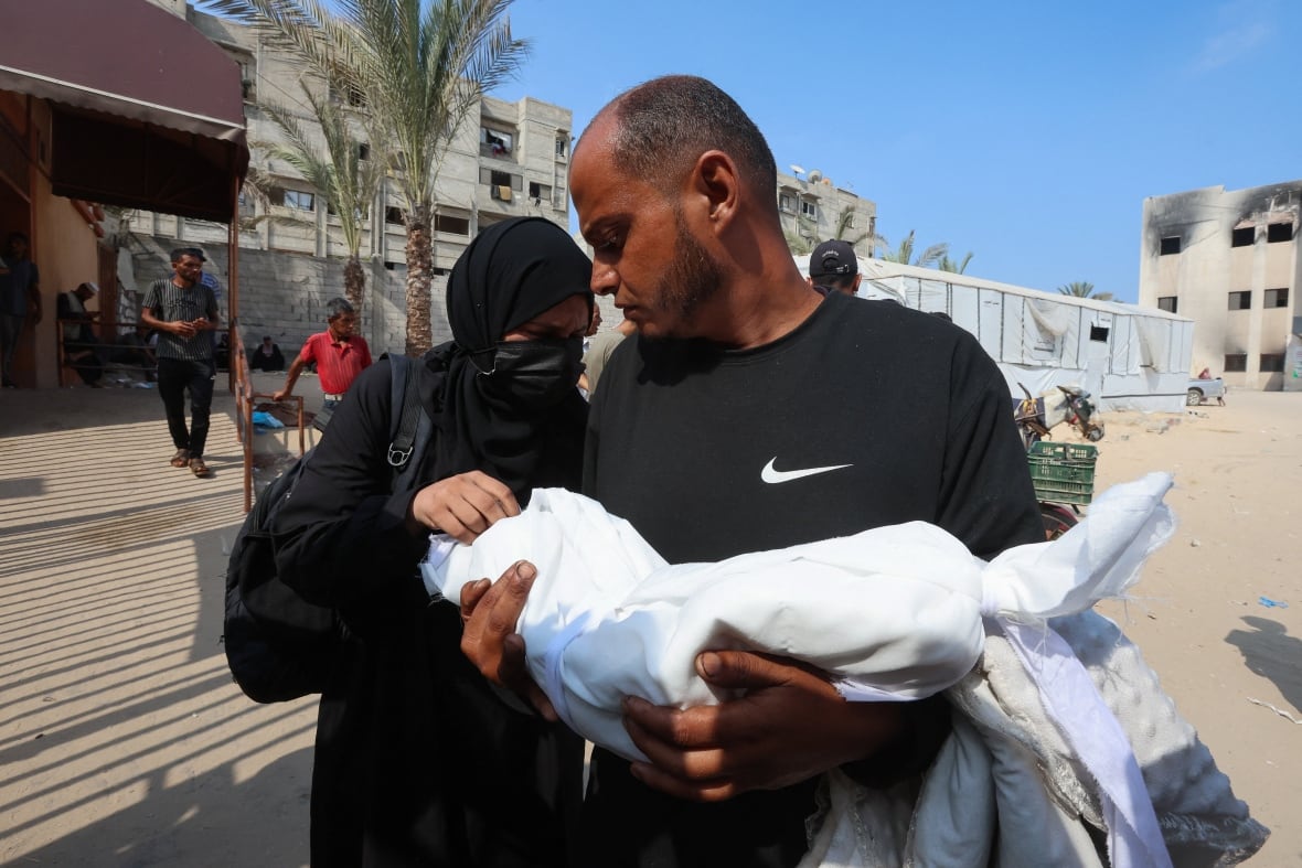 Two people carry the body of a infant wrapped in a bundle of cloth.