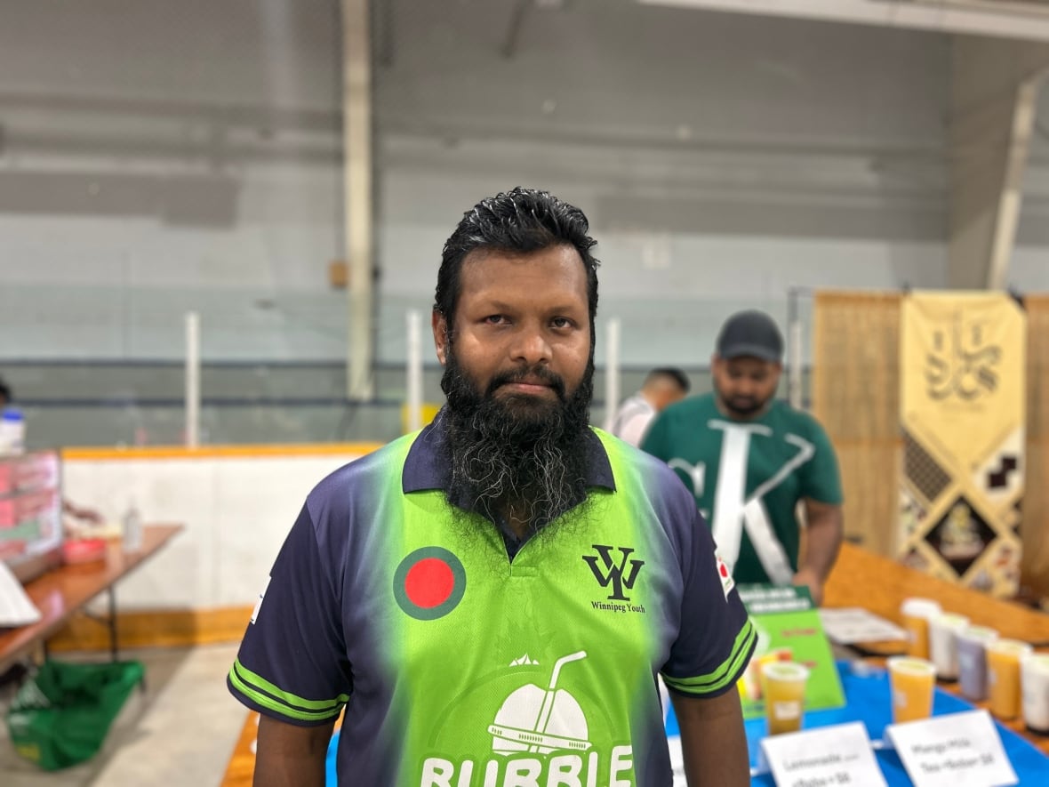 Man with dark hair and a long, dark beard wears a neon green and blue collared shirt, standing in front of a food stall inside an arena