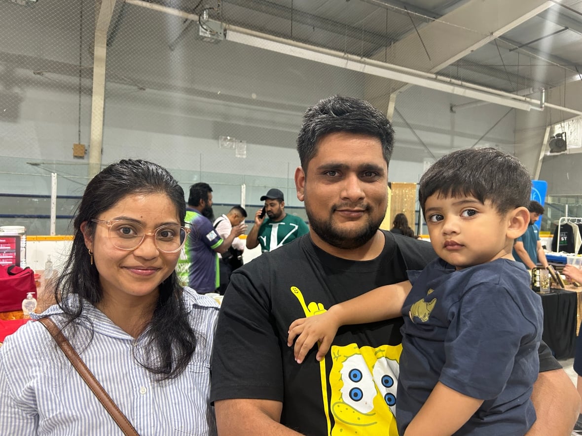 woman with long dark hair and glasses stands next to a man with dark hair and a beard, who is holding a young child with dark hair 
