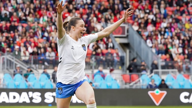 A female soccer player raises both arms in celebration during a match as fans watch from the stands.