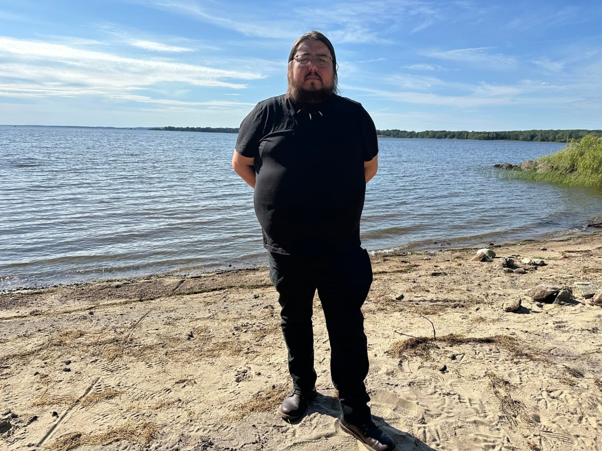 A man with a beard and glasses wearing a black shirt and black pants stands on the sandy shoreline of a lake. The sky is blue.