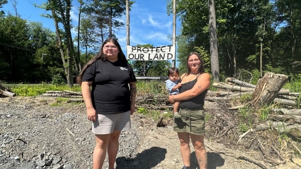 Two young women in t-shirts and shorts stand by a sign that reads, "Protect our Land." They are surrounded by deadfall. The woman on the right is holding a 1 year-old boy.