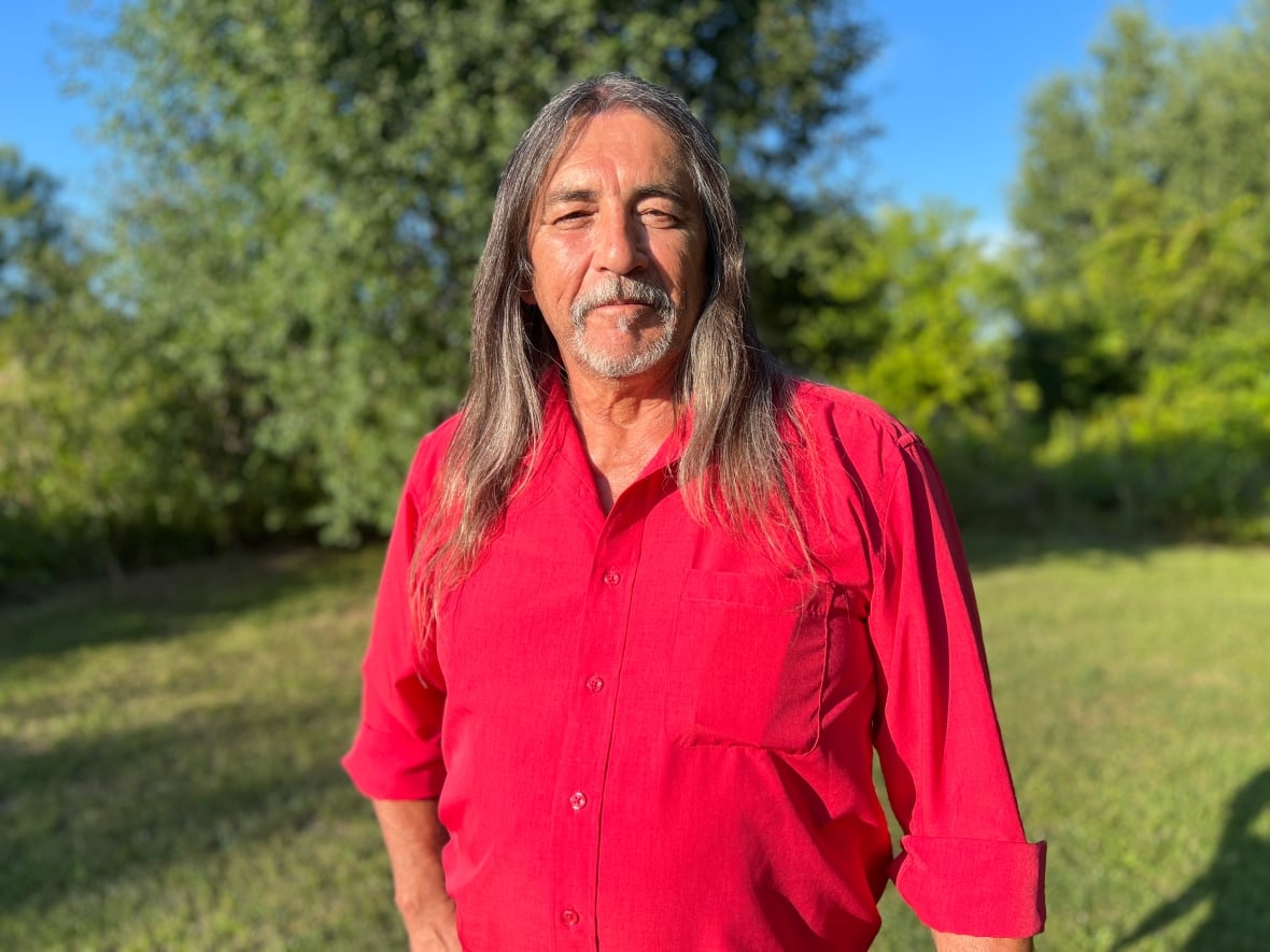 A man with a beard and long, greying hair stands facing the camera wearing a red shirt.
