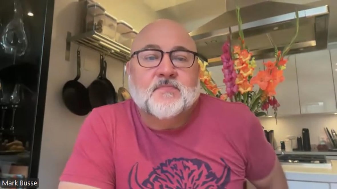 A man speaks in a kitchen wearing a pink shirt.