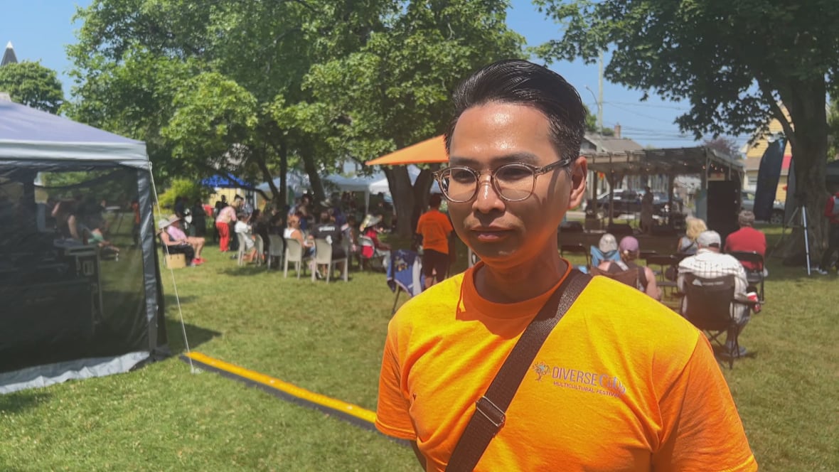 A man wearing glasses and a neon orange shirt. Tents and people sitting in chairs can be seen behind him.