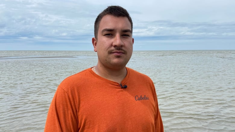 Man with short dark hair wears a long-sleeved orange shirt and stands on a beach beside the water on an overcast day.