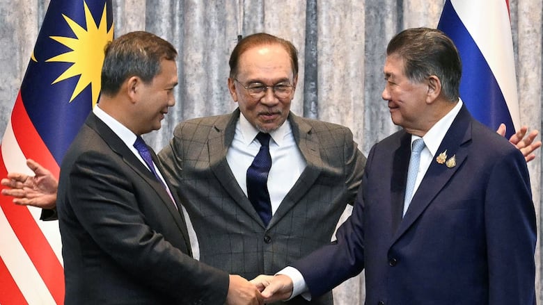 Two men in suits shake hands while a third, between them, extends his arms. 