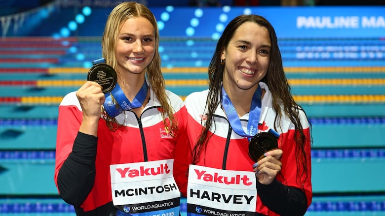 Two female swimmers representing Canada smile while holding up medals near a pool.