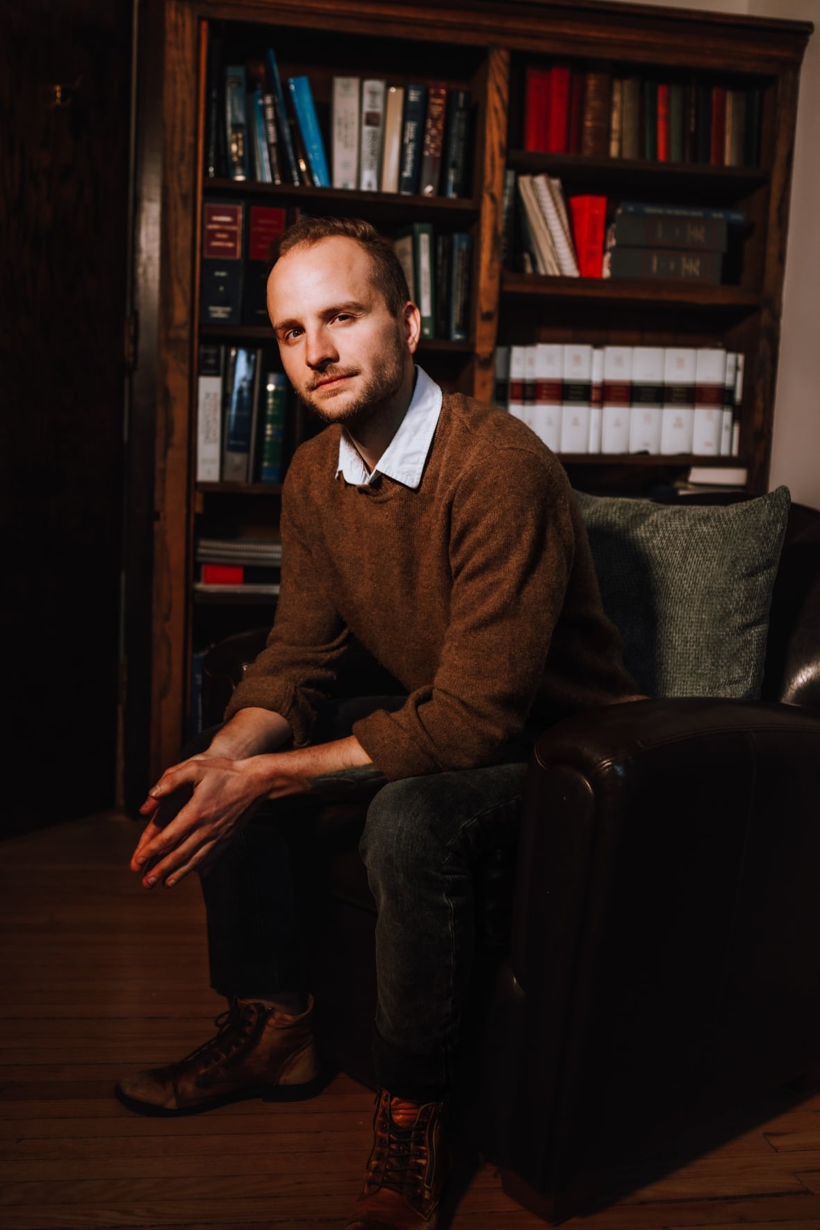 A man in a brown sweater over a white collared t-shirt sits on a chair