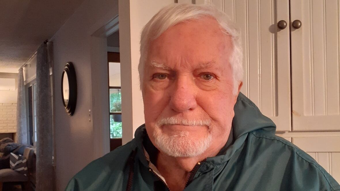 A smiling man with white hair and a goatee in a gray rain jacket sitting in a living room.