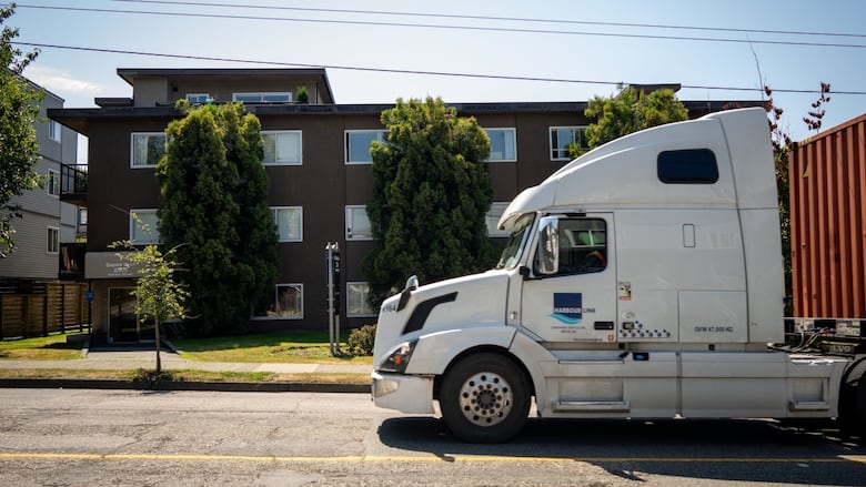 A large white truck drives by a small brown apartment building on a sunny day.