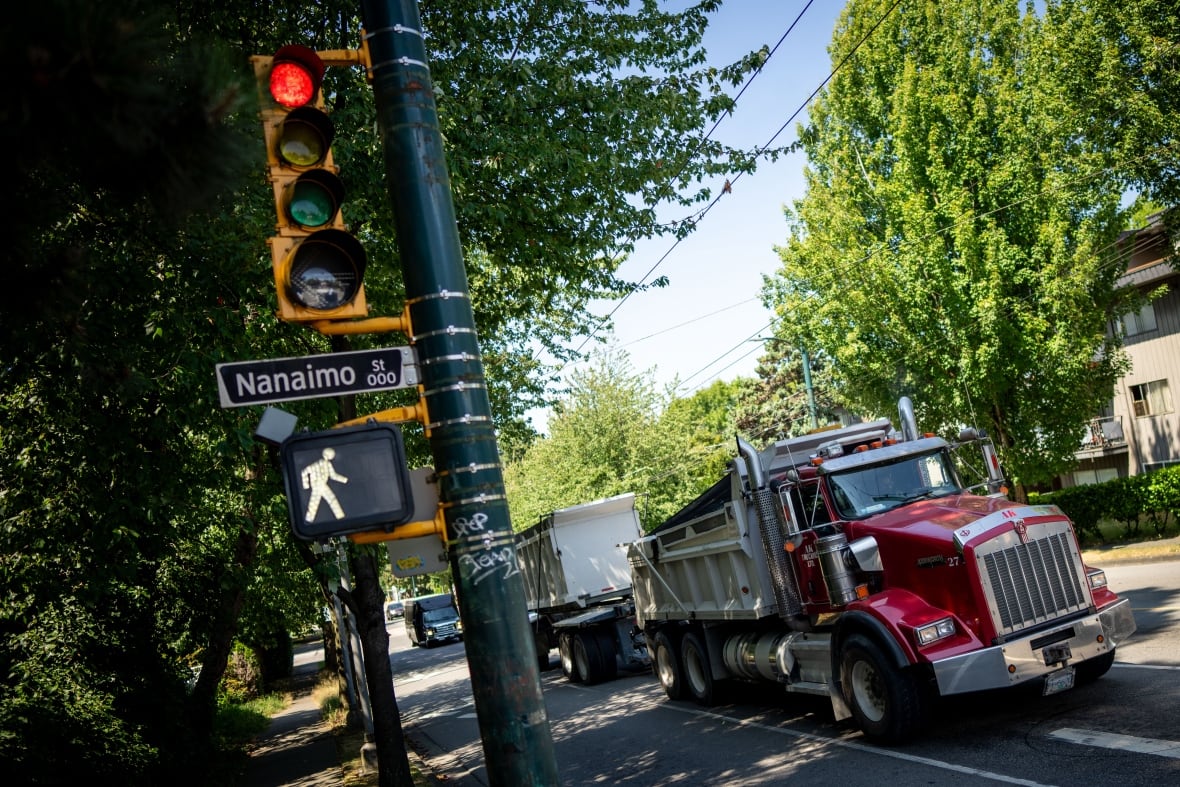  A crosswalk sign is pictured as a large red truck drives down a tree lined street on a sunny day.