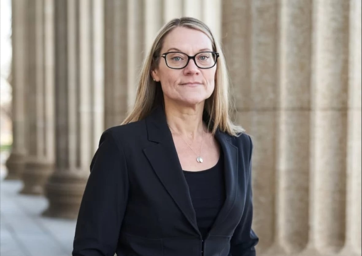 A woman in a black suit and glasses stands in front of large pillars.