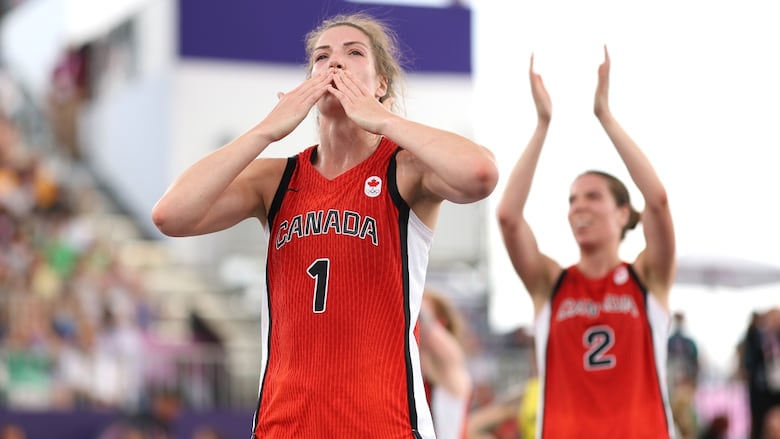 A female three by three basketball player wearing number one blows a kiss to the crowd at a game as teammate claps behind her.