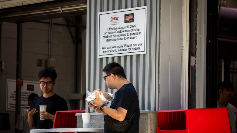 People stand around  eating hot dogs next to a  sign that says: Effective August 5th 2025, an active Costco membership card will be required to purchase items from food court.