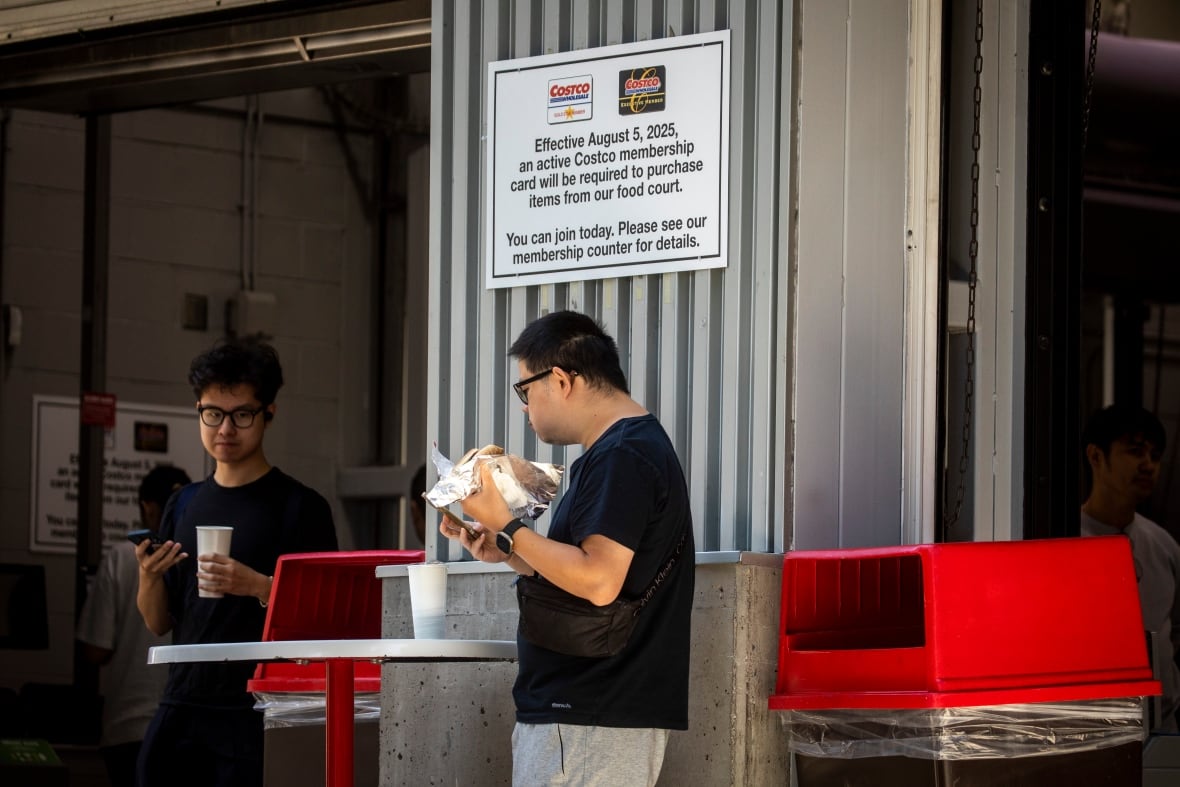 People stand around  eating hot dogs next to a  sign that says: Effective August 5th 2025, an active Costco membership card will be required to purchase items from food court.