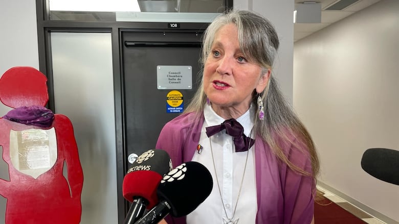 A woman with long grey hair wearing a white dress speaks to reporters.