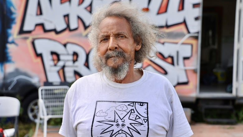 A Black man with grey hair and a beard stands outside a trailer with the words 'Africville protest' in a spray paint design on the side