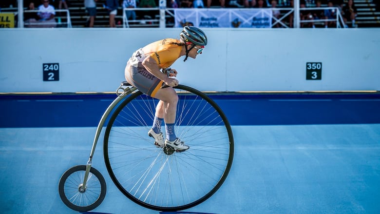 A woman in a yellow cycling suit, bent forward, pedaling a penny farthing on a blue racetrack.  