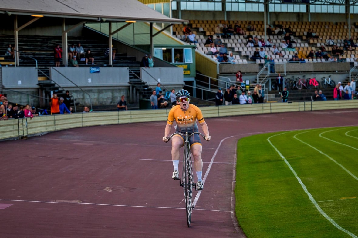 A woman, pictured from the front with her mouth wide open in scream as she rides a penny farthing on a racetrack.