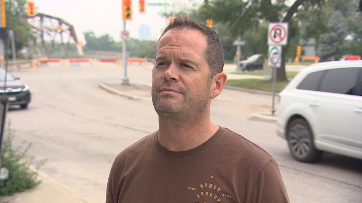 A man with short hair in a brown shirt speaks with a reporter at a busy street corner under construction.