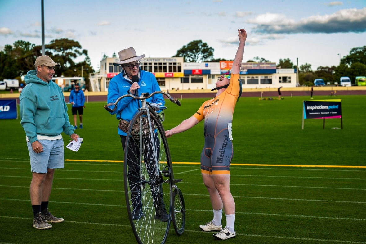 A woman on a racetrack stands next to a penny farthing that's almost as tall as she is. She's holding the bike upright with one hand and throwing the other fist into the air in celebration. 