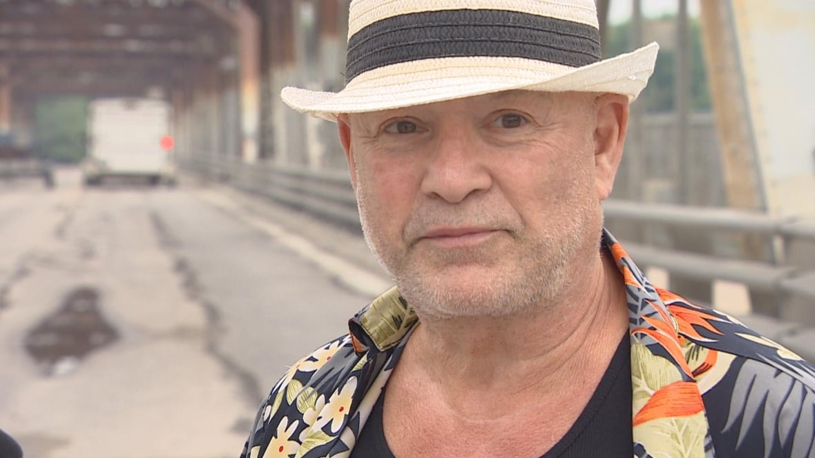 A man wearing a white and black fedora in a shirt with flowers and other patterns stands in front of a bridge.