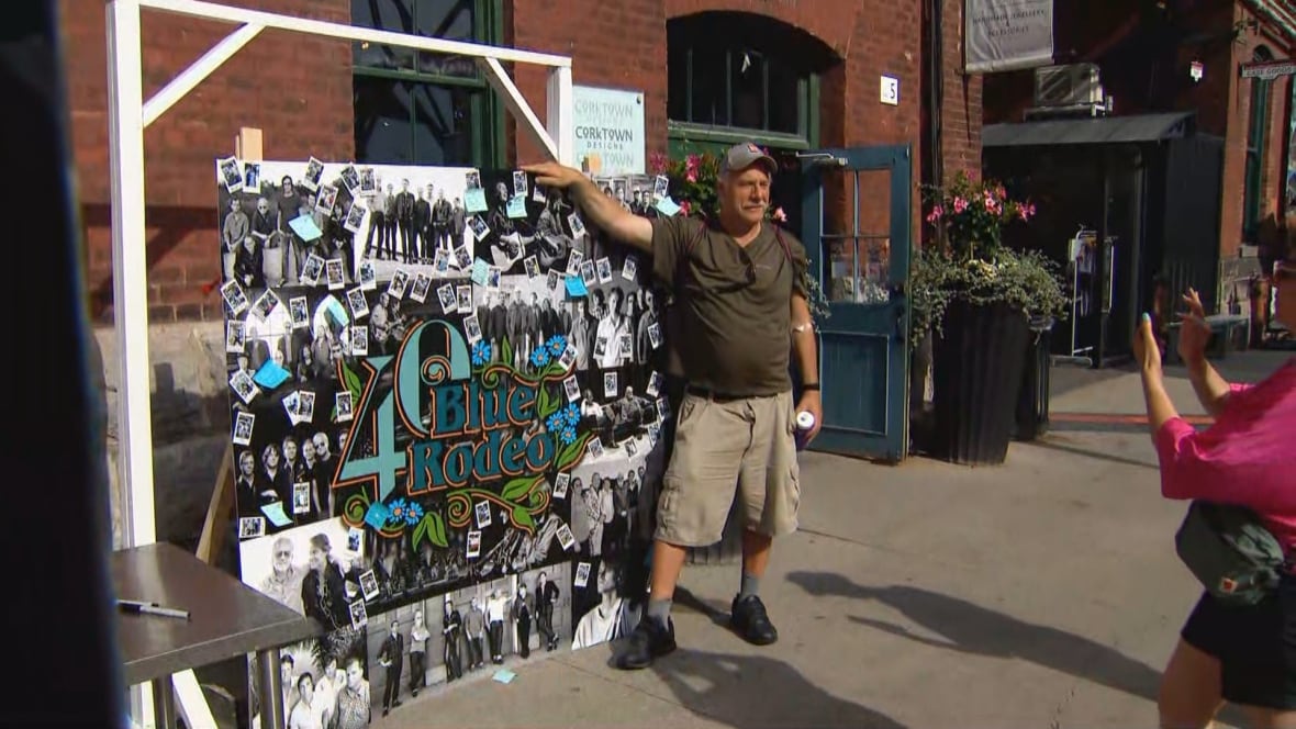 A middle aged man poses for a picture against a collage of Blue Rodeo photos entitled, Blue Rodeo 40, outside a brick building in Toronto's Distillery District on a sunny day