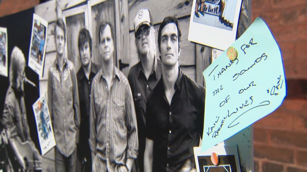 A black and white photo of five members of Blue Rodeo on a poster board. Pinned to it is a post it note that reads: Thanks for the sounds of our lives, signed Paul and Kaili
