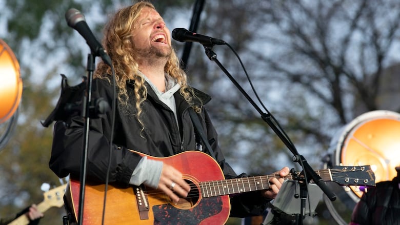  A man with long, blond, curly hair sings at a microphone while playing guitar on a stage outside in autumn. 