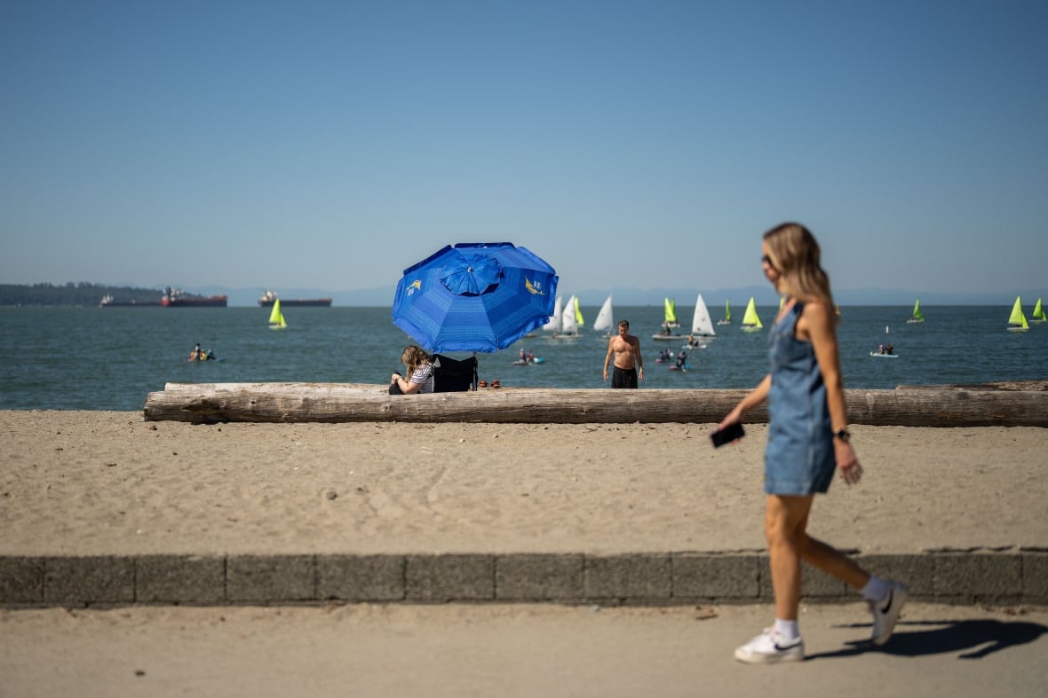 Una mujer camina por una playa con paraguas, botes y gente.