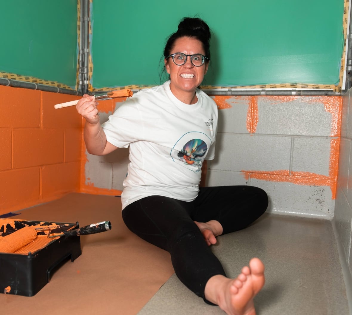A woman sits on the floor with a paint brush and a paint tray. 