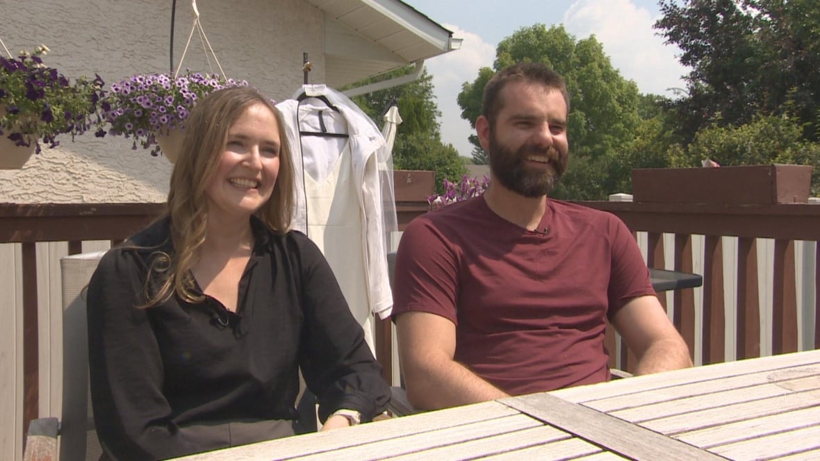 Kelsey and Scot sit outside at a wooden table with a white wedding dress hanging behind them.