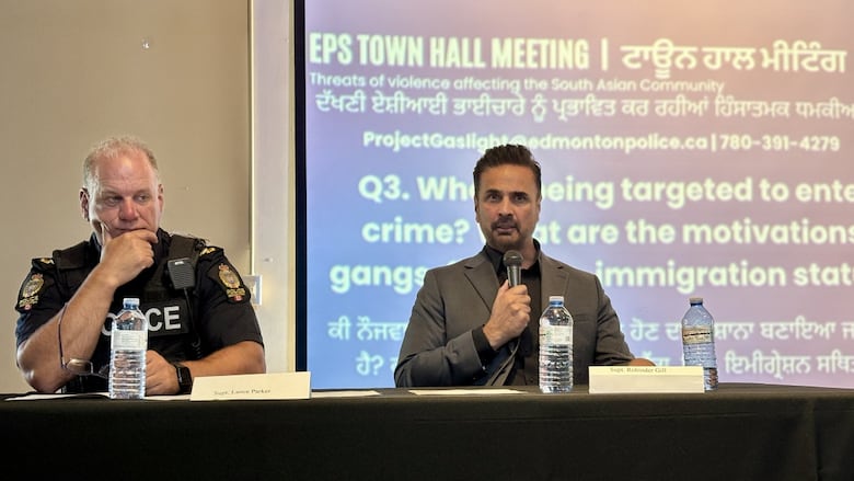 A police officer sits on the left-hand side of the table, with his hand on his chin. He appears to be listening to someone. A man to his right is holding a microphone and looking into a crowd.