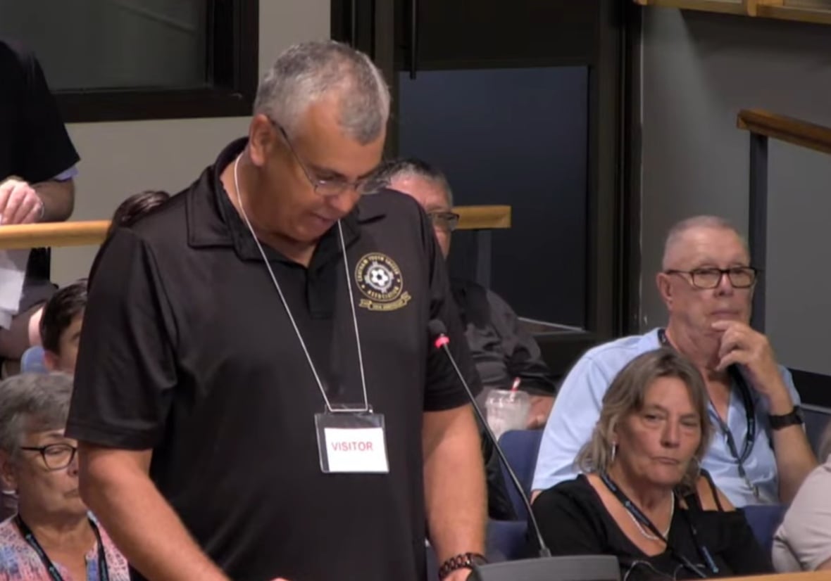 John speaking at the podium with spectators seated behind him.