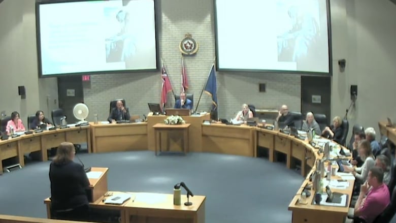 Councilors seated around a horseshoe-shaped series of connected desks. Kim Crew is standing at a podium in front of them while two large screens hang about them displaying her presentation.