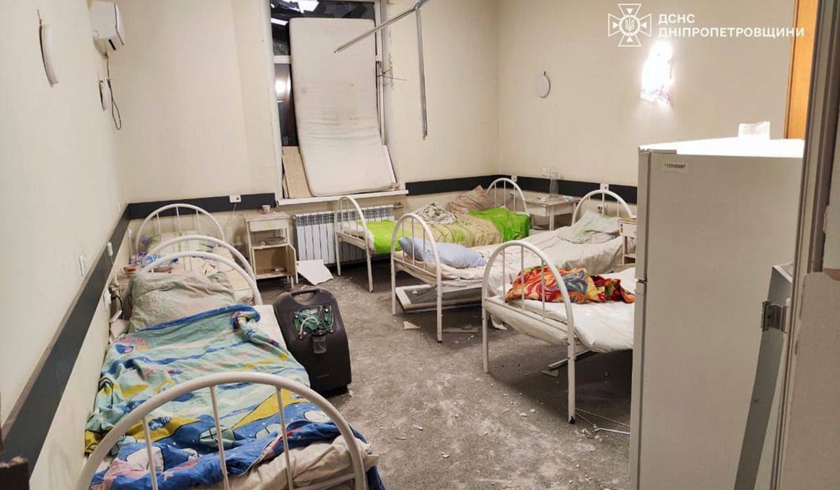 A small room filled with empty hospital beds is shown. There is dust and debris on the floor.