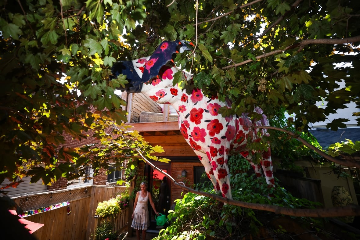 Photo showing a tall yellow papier mache moose with red poppies on it standing outside a home in Toronto surrounded by foliage. There is a woman standing on the entrance to the home.