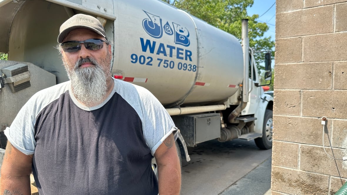 A man wearing sunglasses and a ball cap is shown by a water truck.