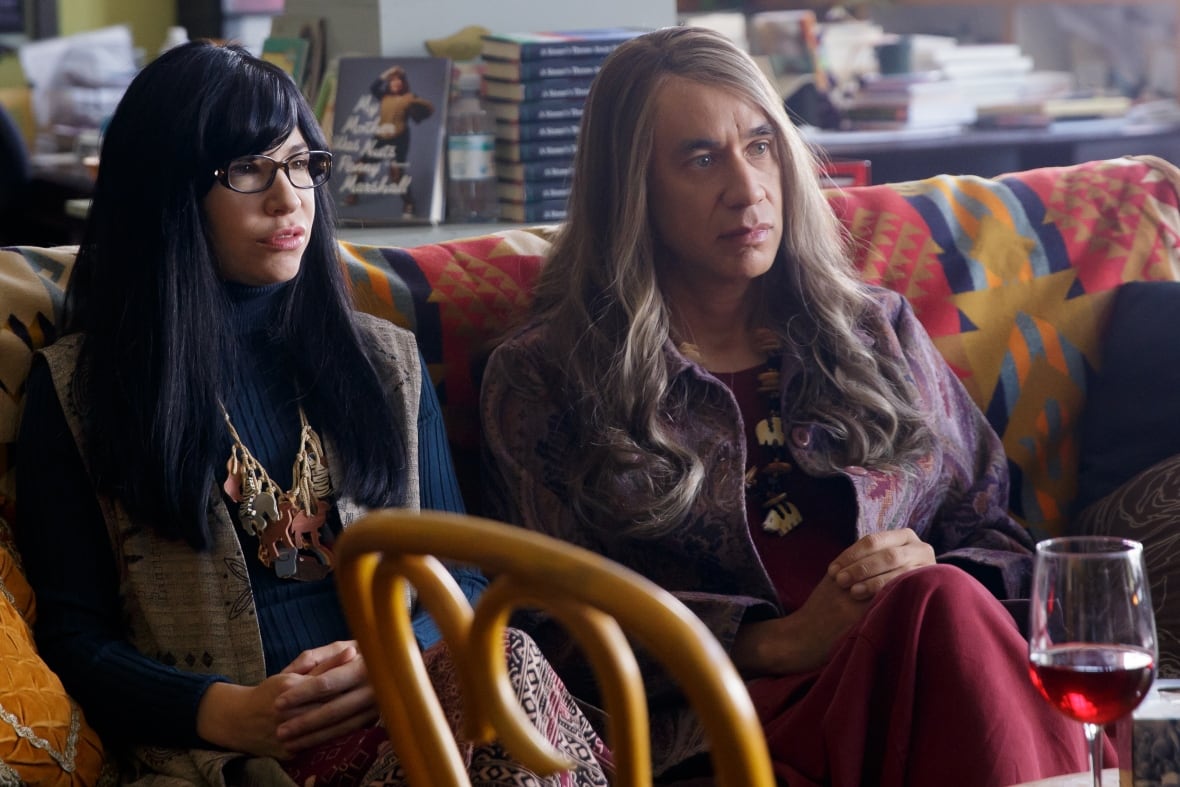 Two people sitting on a couch, one with long black hair and the other with long blonde hair. They appear to be watching television. A glass of wine is in the foreground.